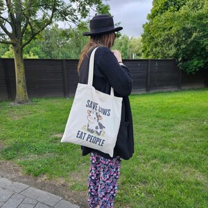 K&ouml;nnte beinhalten: Eine cremefarbene Tragetasche mit dem Text "SAVE COWS EAT PEOPLE" und einer Cartoon-Kuh-Illustration. Die Tasche wird &uuml;ber der Schulter getragen. Die Person tr&auml;gt einen schwarzen Hut und eine Strickjacke.
