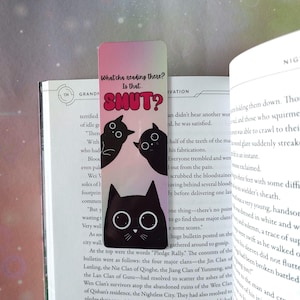 Könnte beinhalten: Schwarzer Katzen-Lesezeichen mit großem weißen Augen und dem Text "What'cha reading there? Is that SMUT?" in Rosa und Weiß. Das Lesezeichen befindet sich in einem Buch mit Text.