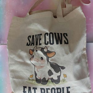 K&ouml;nnte beinhalten: Beige Canvas-Tragetasche mit schwarzem Text "SAVE COWS EAT PEOPLE". Die Tasche zeigt eine Cartoon-Kuh-Illustration mit schwarz-wei&szlig;en Flecken und kleinen gelben Blumen.