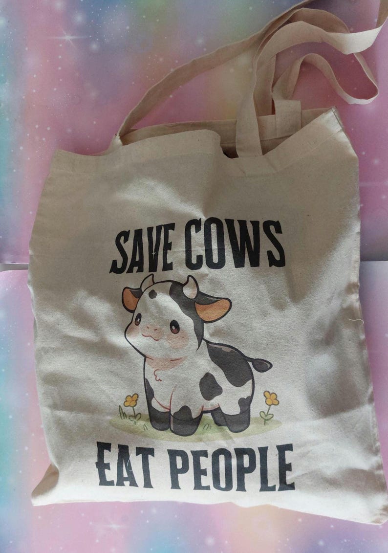 K&ouml;nnte beinhalten: Beige Canvas-Tragetasche mit schwarzem Text "SAVE COWS EAT PEOPLE". Die Tasche zeigt eine Cartoon-Kuh-Illustration mit schwarz-wei&szlig;en Flecken und kleinen gelben Blumen.
