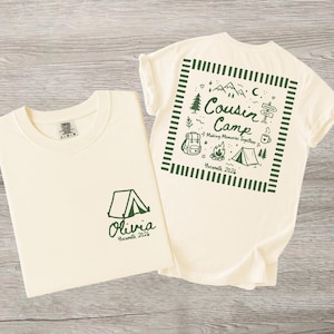 May include: A cream-coloured t-shirt featuring dark green graphics. The front displays a tent icon with the text 'Olivia Yosemite 2026'. The back shows a square frame containing a tent, campfire, backpack, mountains, and the text 'Cousin Camp Making Memories Together Yosemite 2026'.