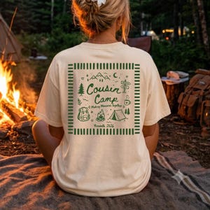 May include: A cream-coloured t-shirt featuring a dark green graphic on the back. The design includes the text "Cousin Camp Making Memories Together Yosemite 2026" surrounded by illustrations of mountains, a tent, a campfire, and a backpack.