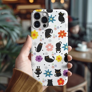 May include: White phone case featuring a repeating pattern of black cat illustrations and colorful flowers. The case has a glossy finish and a camera cutout. The design includes yellow, pink, blue, and purple flowers, and small white flower accents.