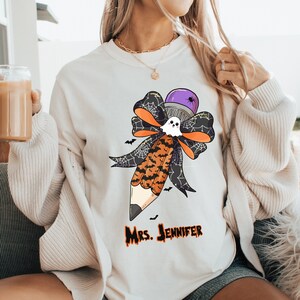 Pu&ograve; includere: Felpa color crema con un design a tema Halloween a forma di matita. La matita &egrave; decorata con un fantasma, pipistrelli e un fiocco. Il testo "Mrs. Jennifer" &egrave; stampato in arancione sotto il disegno. La felpa ha un girocollo.