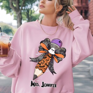 Pu&ograve; includere: Una felpa rosa chiaro con un disegno a matita a tema Halloween. La matita ha un fiocco con ragnatele e pipistrelli, un fantasma e la scritta "Mrs. Jennifer". La matita &egrave; decorata con pipistrelli e zucche.