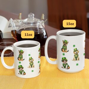 May include: Two white ceramic mugs with handles, one 11oz and one 15oz, featuring a repeating pattern of dogs wearing green leprechaun hats and holding shamrocks. The mugs are filled with dark liquid and are on a yellow and green placemat.