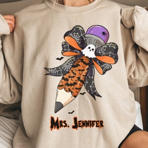 Pu&ograve; includere: Felpa beige con un disegno a matita a tema Halloween. La matita presenta una gomma viola, un fantasma e un fiocco con ragnatele e pipistrelli. Il testo "Mrs. Jennifer" &egrave; stampato in arancione e nero.