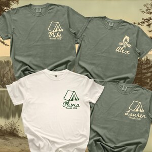 May include: Three sage green t-shirts and one white t-shirt, each featuring a small graphic on the left chest. The graphics include a tent or campfire icon, a name in script font, and the text Yosemite 2026 printed in a simple sans-serif font.