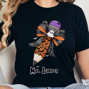 Pu&ograve; includere: T-shirt nera con un disegno a matita a tema Halloween. La matita ha una gomma a forma di fantasma, una parte superiore viola con un ragno e un fiocco con accenti arancioni e neri. Il testo "Mrs. Jennifer" &egrave; stampato sotto.