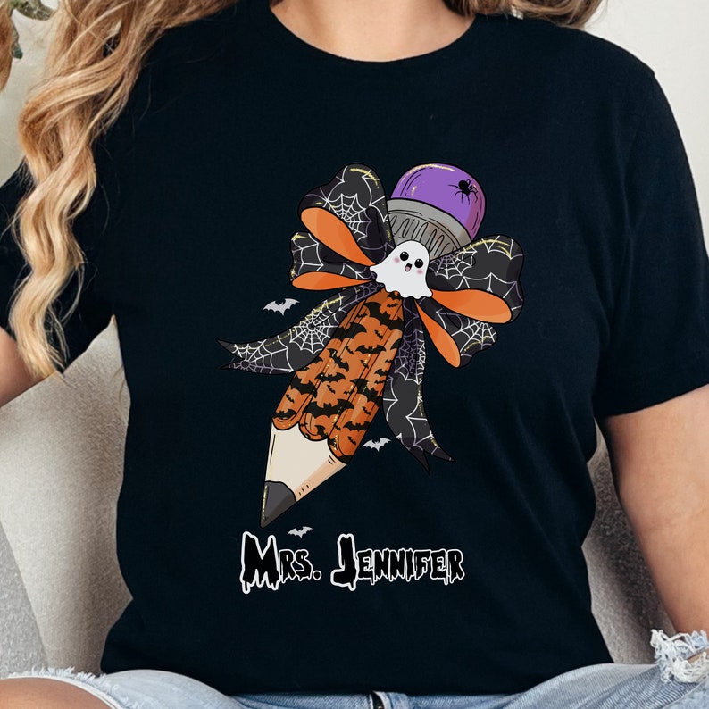 Pu&ograve; includere: T-shirt nera con un disegno a matita a tema Halloween. La matita ha una gomma a forma di fantasma, una parte superiore viola con un ragno e un fiocco con accenti arancioni e neri. Il testo "Mrs. Jennifer" &egrave; stampato sotto.