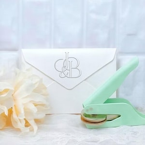 May include: A mint green handheld embosser tool with a metallic circular die, positioned next to a white envelope featuring a silver embossed monogram of the letters J and B. The tool has a lever mechanism for pressing designs onto paper stationery.
