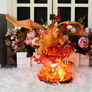 May include: A light-up figurine of a Charizard Pokemon, with a red and orange body, and a yellow and orange tail. The figurine is surrounded by flames and has a small Charmander and a small Charmeleon figurine next to it.