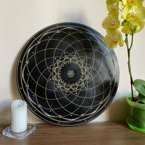 May include: A round, black decorative wall art piece with a geometric design in gold and white. The artwork features a central star pattern and a series of concentric circles. A white candle and yellow orchid are in the background.