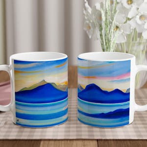 May include: Two white ceramic mugs with a blue and green mountain landscape design. The mugs have a white handle and are on a checkered tablecloth.