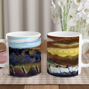 May include: Two white ceramic mugs with handles. The mug on the left has a painting of a landscape with a blue sky, green hills, and a fence. The mug on the right has a painting of a field with sheep walking through it.