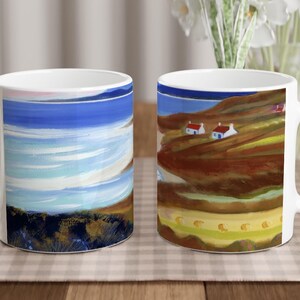 May include: Two white ceramic mugs with colorful painted designs. One mug features a blue and white seascape with a sandy beach. The other mug features a brown and green landscape with white houses.
