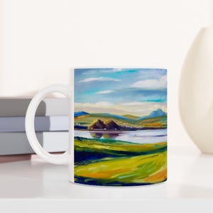 May include: White ceramic mug with a colorful landscape scene printed on it. The scene features a lake, hills, and a cloudy sky.
