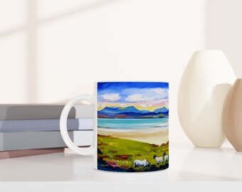 May include: White ceramic mug with a colorful painting of a beach scene with mountains in the background. The painting features a blue sky, white clouds, a turquoise ocean, a sandy beach, and green grass. Three white sheep are in the foreground.