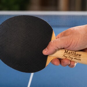 May include: A black ping pong paddle with a wooden handle is held in a person's hand. The handle is engraved with the name "Matthew" and the words "Ping Pong Master". The paddle has a textured rubber surface.