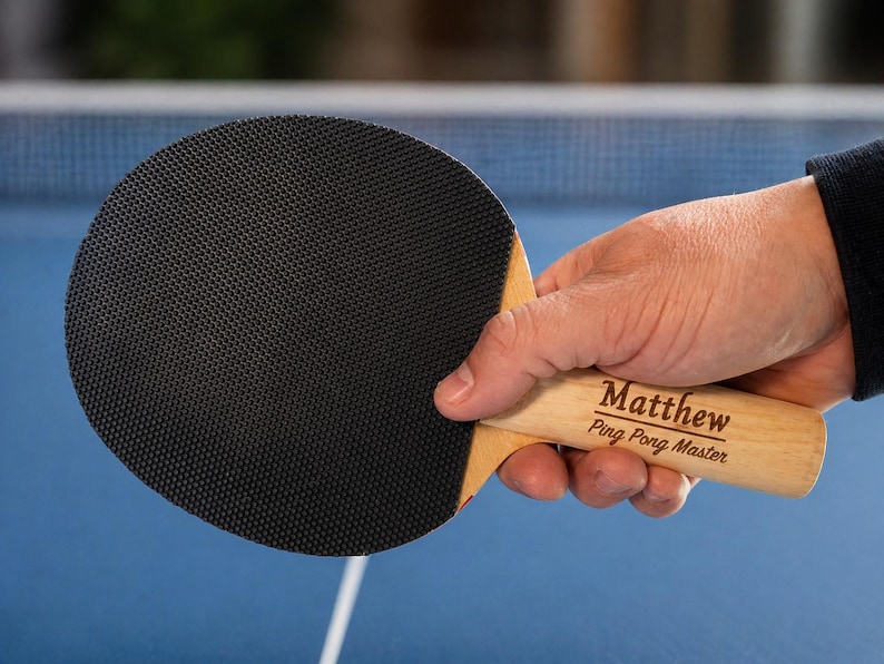 May include: A black ping pong paddle with a wooden handle is held in a person's hand. The handle is engraved with the name "Matthew" and the words "Ping Pong Master". The paddle has a textured rubber surface.