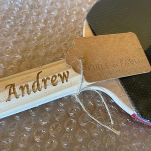 Puede incluir: Una raqueta de ping pong de madera con una superficie negra y un borde rojo. El mango de la raqueta est&aacute; grabado con el nombre "Andrew". Una etiqueta marr&oacute;n est&aacute; adjunta con el texto "FIRLE & FABLE UNIQUE HEARTS".