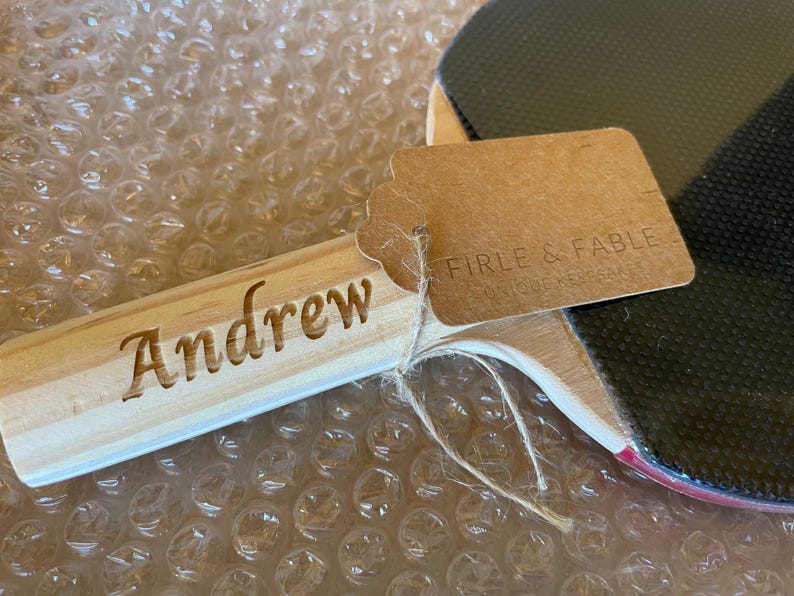 Puede incluir: Una raqueta de ping pong de madera con una superficie negra y un borde rojo. El mango de la raqueta est&aacute; grabado con el nombre "Andrew". Una etiqueta marr&oacute;n est&aacute; adjunta con el texto "FIRLE & FABLE UNIQUE HEARTS".