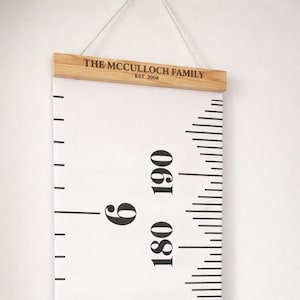 Pode incluir: Um gráfico de altura branco com marcações e números pretos, incluindo 180 e 190 cm. O gráfico é suspenso por uma corda e tem um topo de madeira com o texto "THE MCCULLOCH FAMILY EST. 2008".