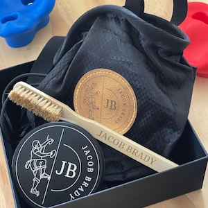 Puede incluir: Una caja de regalo negra contiene una bolsa de magnesio para escalada, un cepillo de madera y dos parches redondos. Los parches y el cepillo están marcados con "JACOB BRADY" y las iniciales "JB". En el fondo se ven presas de escalada azules y rojas.