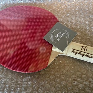 May include: A red ping pong paddle with a wooden handle. The handle is engraved with the name "Eli" and the words "Ping Pong Master". A gray tag with the words "Firle & Fable" is attached to the paddle.
