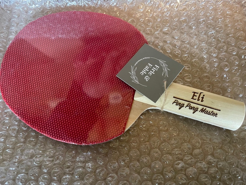 May include: A red ping pong paddle with a wooden handle. The handle is engraved with the name "Eli" and the words "Ping Pong Master". A gray tag with the words "Firle & Fable" is attached to the paddle.