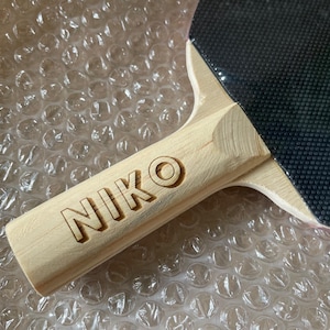 Puede incluir: Primer plano de una raqueta de ping pong con mango de madera. El mango est&aacute; grabado con el nombre "NIKO". La superficie de juego es negra y el mango es de madera clara.