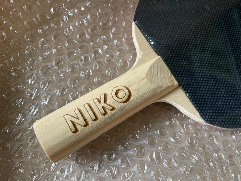 Puede incluir: Primer plano de una raqueta de ping pong con mango de madera. El mango est&aacute; grabado con el nombre "NIKO". La superficie de juego es negra y el mango es de madera clara.