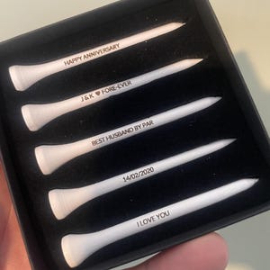 Könnte beinhalten: Ein Set aus fünf weißen Golf-Tees mit schwarzem Text in einer schwarzen Box. Der Text auf den Tees lautet: "HAPPY ANNIVERSARY", "J&K FORE-EVER", "BEST HUSBAND BY PAR", "14/02/2020" und "I LOVE YOU".