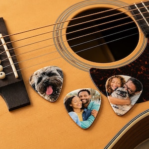 Puede incluir: Tres púas de guitarra personalizadas con impresiones fotográficas sobre el cuerpo de madera clara de una guitarra acústica. Una púa muestra un perro con la lengua fuera, otra una pareja abrazada y la tercera una persona con un perro.