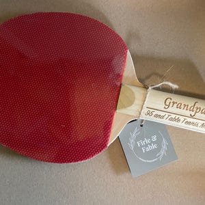 May include: A ping pong paddle with a red rubber surface and a wooden handle. The handle is engraved with the word "Grandpa" and "95 and Table Tennis Master". A gray tag with the words "Firle & Fable" is attached.
