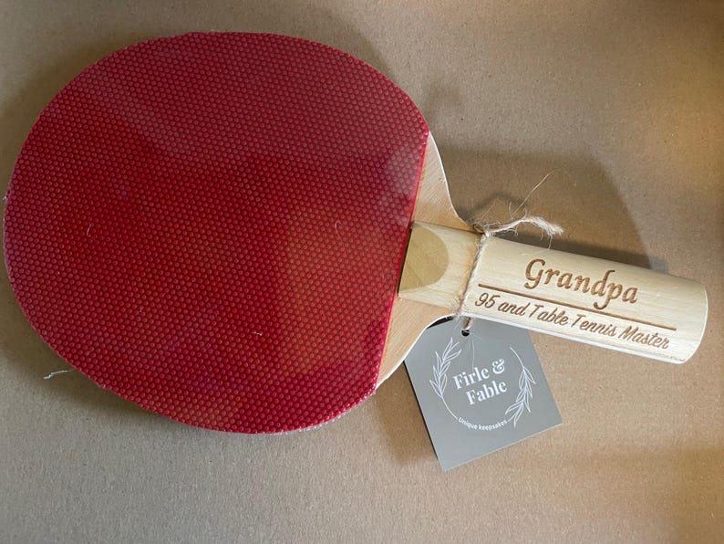 May include: A ping pong paddle with a red rubber surface and a wooden handle. The handle is engraved with the word "Grandpa" and "95 and Table Tennis Master". A gray tag with the words "Firle & Fable" is attached.