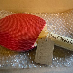 May include: A red ping pong paddle with a wooden handle, featuring the name "Paul Welsh" engraved on the handle. A brown tag is attached with string. The paddle is in a cardboard box with bubble wrap.