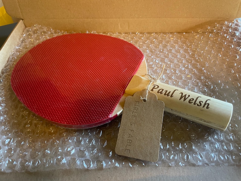 May include: A red ping pong paddle with a wooden handle, featuring the name "Paul Welsh" engraved on the handle. A brown tag is attached with string. The paddle is in a cardboard box with bubble wrap.