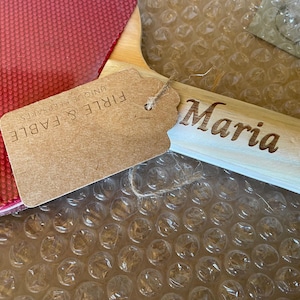 Puede incluir: Una paleta de madera con el nombre "Maria" grabado en el mango. Una etiqueta con el texto "FIRLE & FABLE UNIQUE KEEPSAKES" est&aacute; adjunta. La paleta tiene una superficie roja y est&aacute; sobre pl&aacute;stico de burbujas.