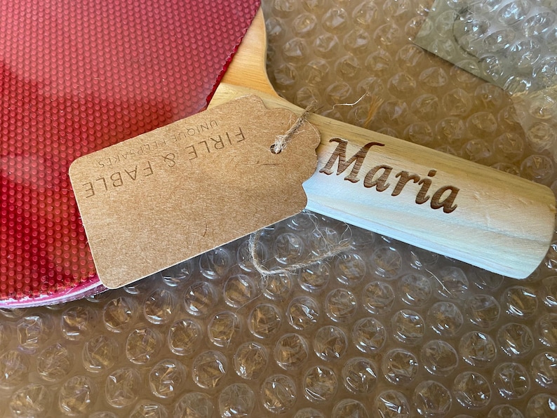 Puede incluir: Una paleta de madera con el nombre "Maria" grabado en el mango. Una etiqueta con el texto "FIRLE & FABLE UNIQUE KEEPSAKES" est&aacute; adjunta. La paleta tiene una superficie roja y est&aacute; sobre pl&aacute;stico de burbujas.