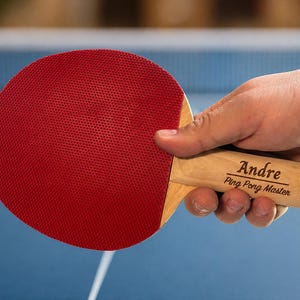 May include: A red table tennis bat with a wooden handle. The handle is engraved with the name "Andre" and the text "Ping Pong Master". The bat is held against a blue background, possibly during a match.