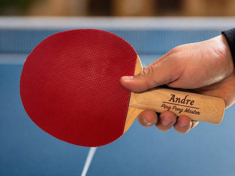 May include: A red ping pong paddle with a wooden handle. The handle is engraved with the name "Andre" and the text "Ping Pong Master". The paddle is held against a blue background, suggesting a game in progress.