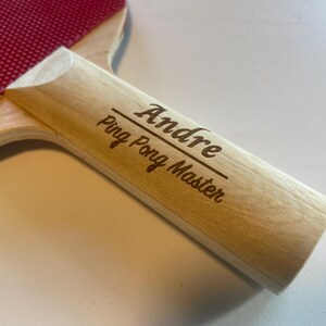 May include: A ping pong paddle with a red rubber surface and a light wood handle. The handle is engraved with the name "Andre" above the words "Ping Pong Master".