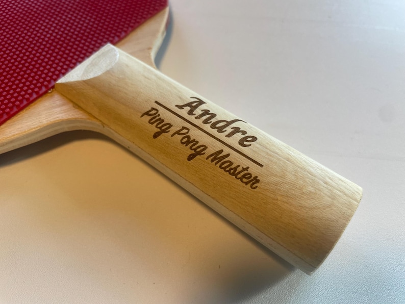 May include: A ping pong paddle with a red rubber surface and a light wood handle. The handle is engraved with the name "Andre" above the words "Ping Pong Master".
