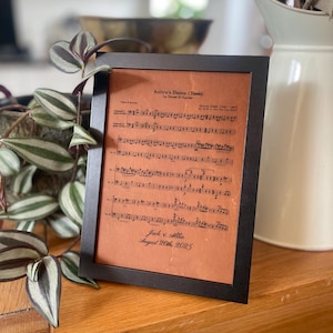 May include: A framed print featuring sheet music titled "Anitra's Dance (Tema)" in a black frame. The print has a warm, sepia-toned background with musical notes and text. The names "Jack & Allie" and the date "August 20th, 2025" are also visible.