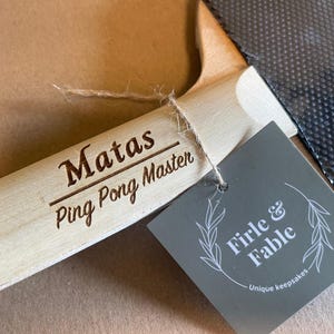 May include: A light-colored wooden ping pong paddle handle with the engraved text "Matas Ping Pong Master." A gray tag with the text "Firle & Fable" and a leaf design is attached. The paddle's black surface is visible.