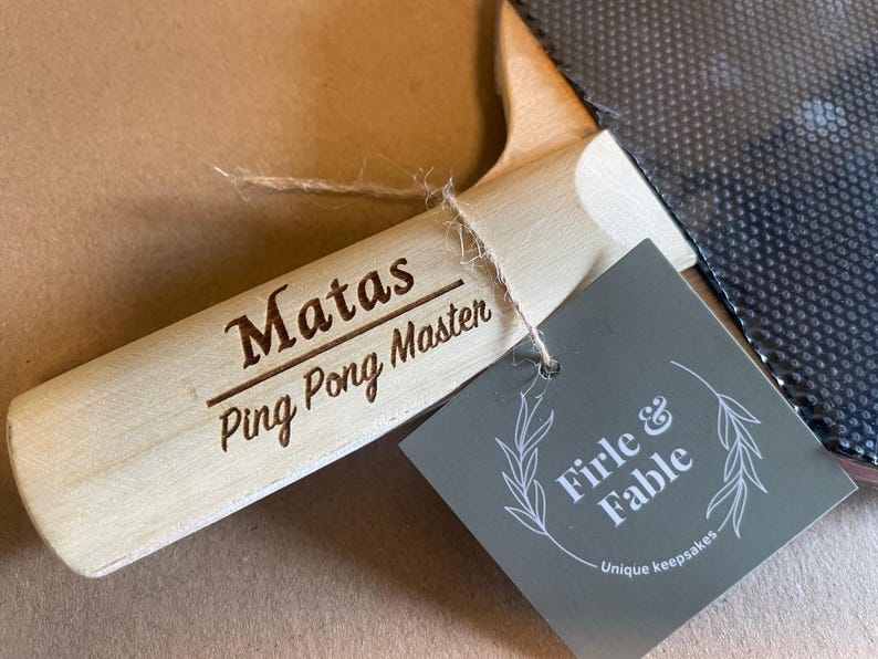 May include: A light-colored wooden ping pong paddle handle with the engraved text "Matas Ping Pong Master." A gray tag with the text "Firle & Fable" and a leaf design is attached. The paddle's black surface is visible.