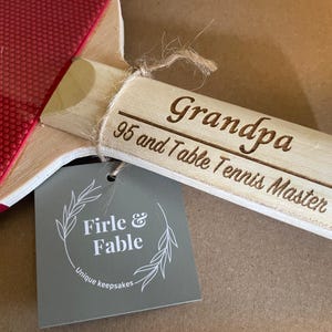 May include: A wooden table tennis paddle with a red rubber surface and a wooden handle. The handle is engraved with "Grandpa 95 and Table Tennis Master." A gray tag reads "Firle & Fable, Unique keepsakes."