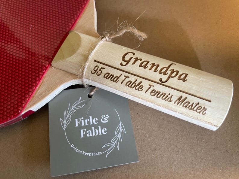 May include: A wooden table tennis paddle with a red rubber surface and a wooden handle. The handle is engraved with "Grandpa 95 and Table Tennis Master." A gray tag reads "Firle & Fable, Unique keepsakes."