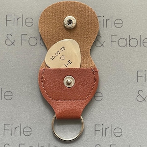 Könnte beinhalten: Ein brauner Gitarrenplektrumhalter aus Leder mit einem silbernen Schlüsselring. Im Inneren ist ein silbernes Gitarrenplektrum zu sehen, graviert mit dem Datum "10.07.23", einem Herzsymbol und den Initialen "J+E". Der Halter ist offen.